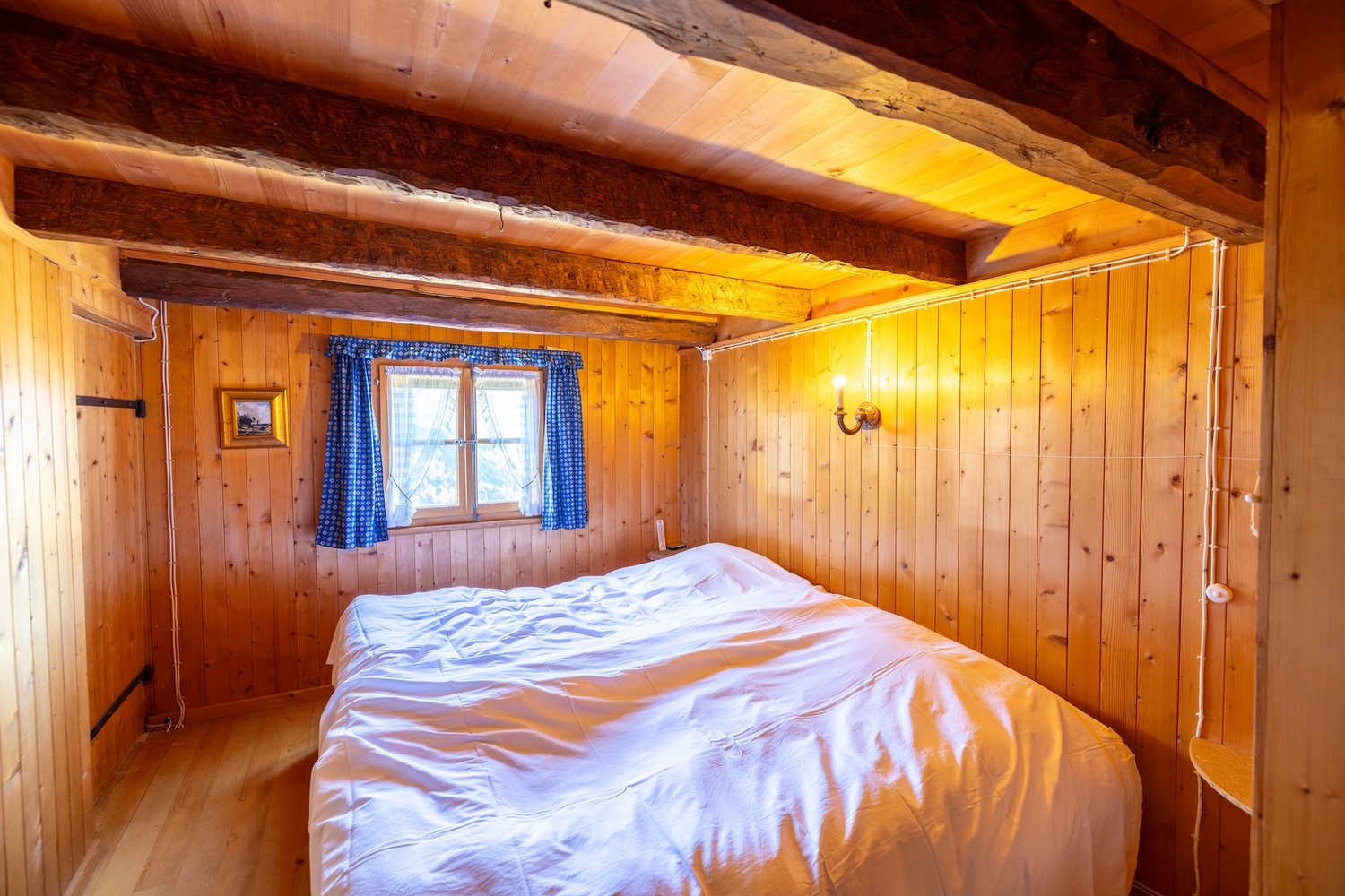 Chambre lambrissée du mayen : lit double sous les poutres, petite fenêtre à rideaux bleus sur la montagne.