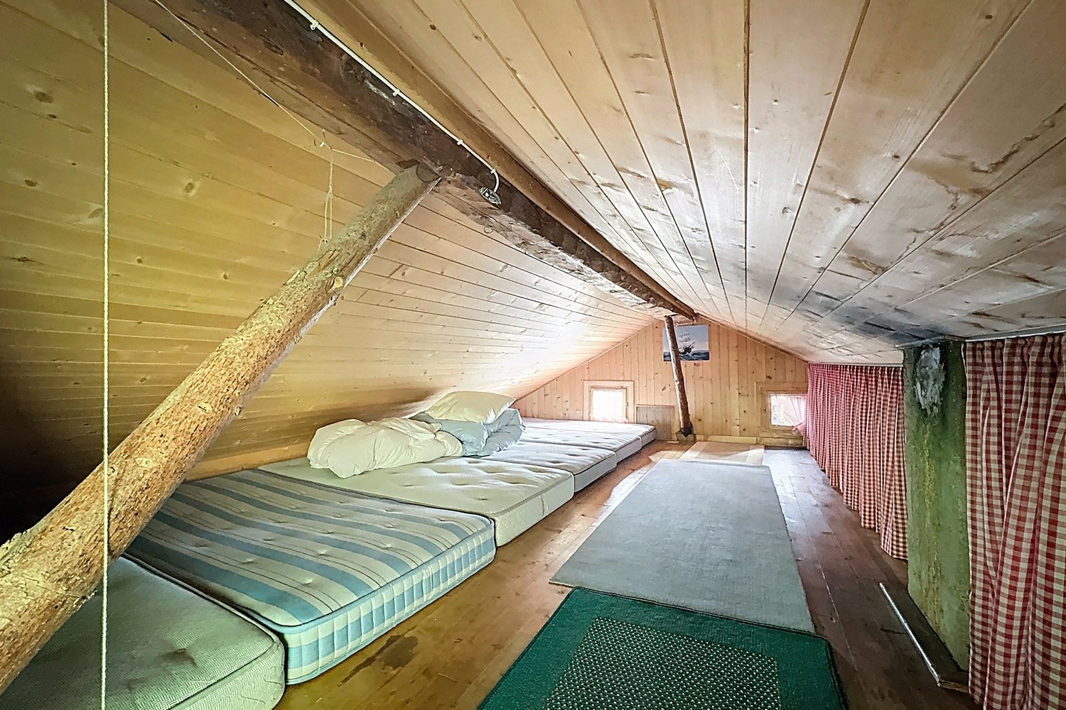 Dortoir sous les combles du mayen : plafond en planches claires, matelas alignés au sol, rideau à carreaux rouges.