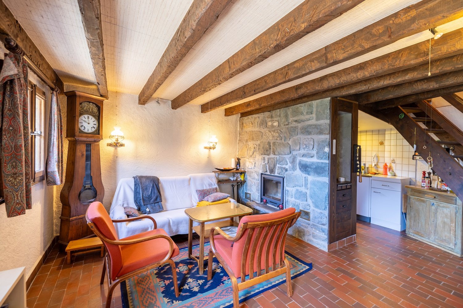 Séjour du mayen : fauteuils autour d'une table basse, tomettes, poutres apparentes, cheminée en pierre avec insert à bois.