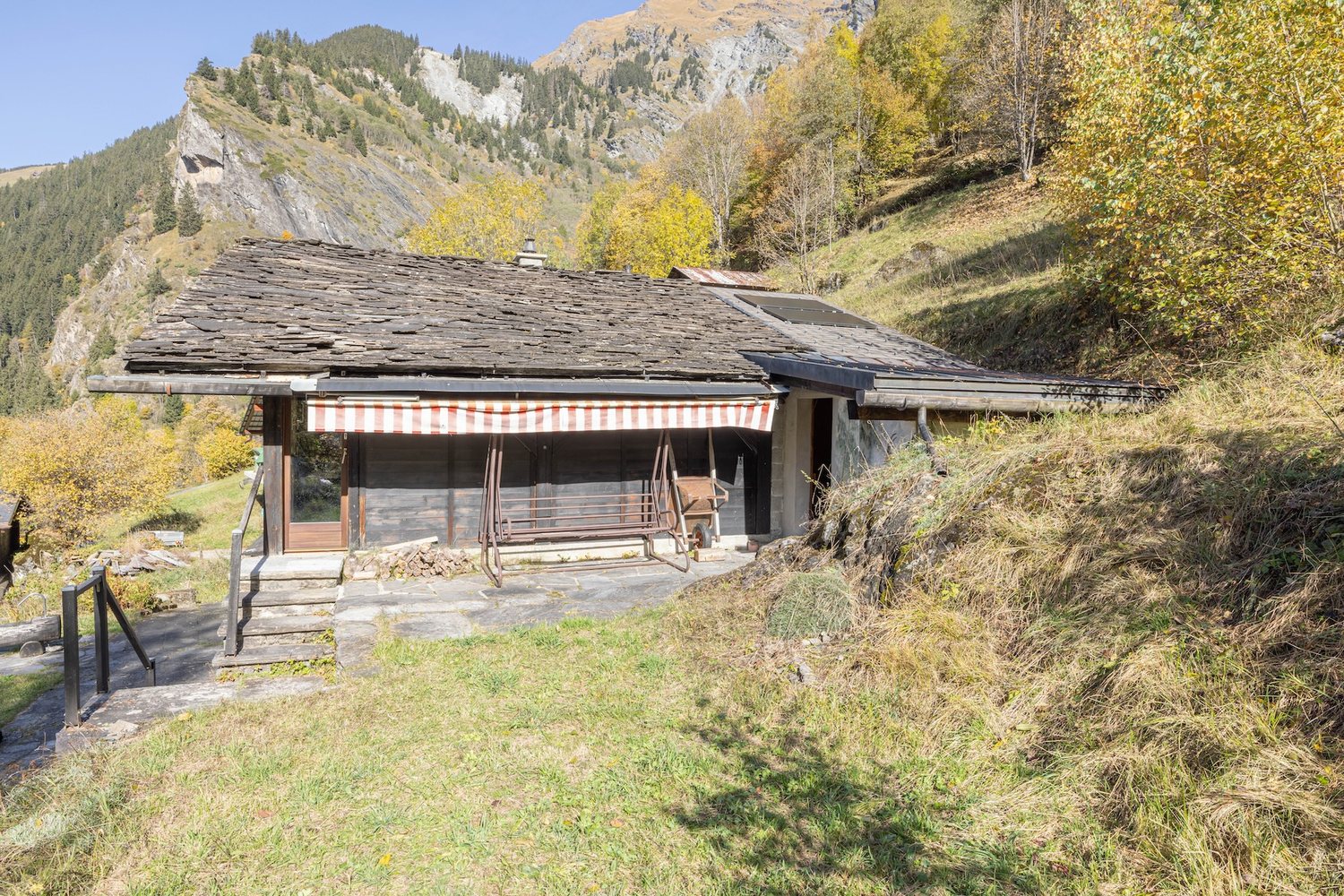 Le toit en dalles de pierre du mayen vu depuis la pente, terrasse haute abritée par un store rayé, mélèzes et rochers derrière.