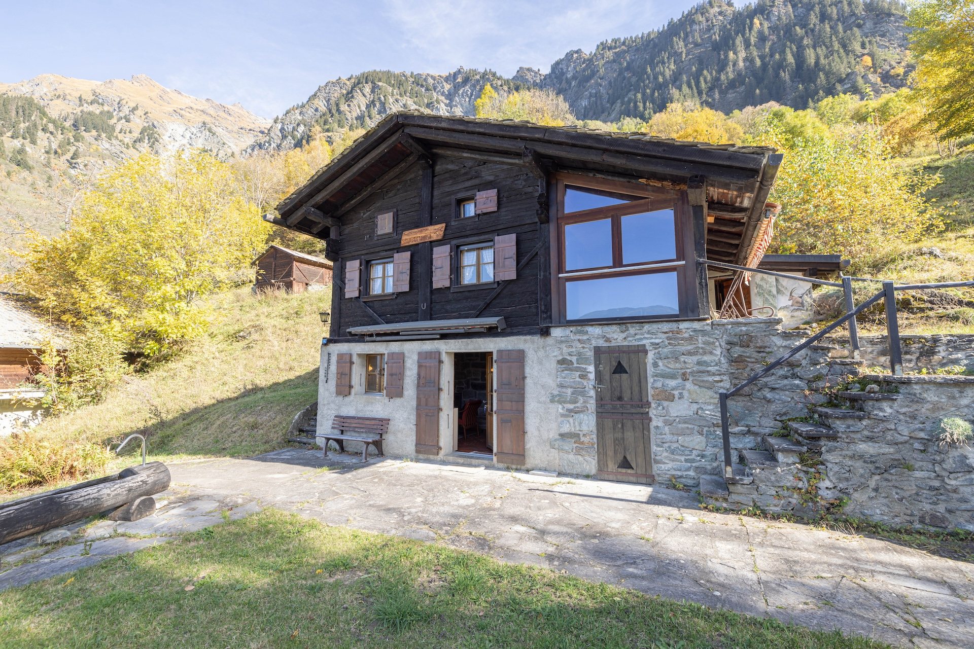 Le Mayen des Planches à Lourtier : chalet en madriers sombres sur socle de pierre, pelouse et bassin en mélèze au premier plan, mélèzes dorés et montagnes du Haut Val de Bagnes derrière.