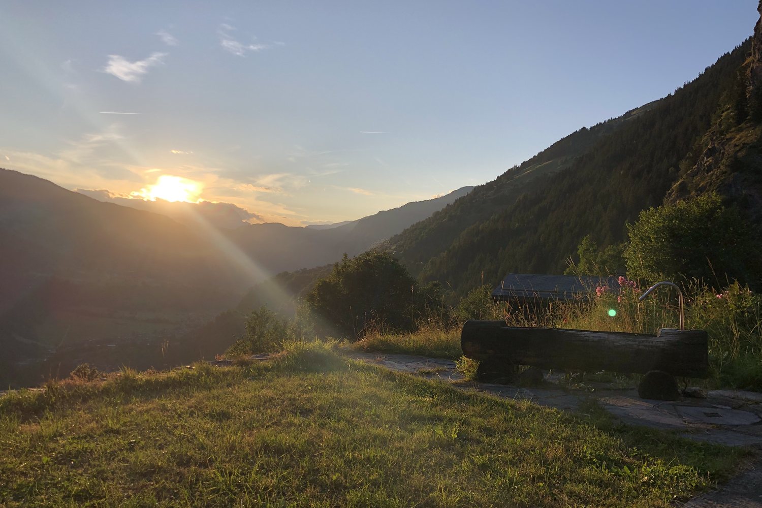 Coucher de soleil depuis la terrasse du mayen : le soleil descend derrière les crêtes, la fontaine en mélèze en contre-jour.